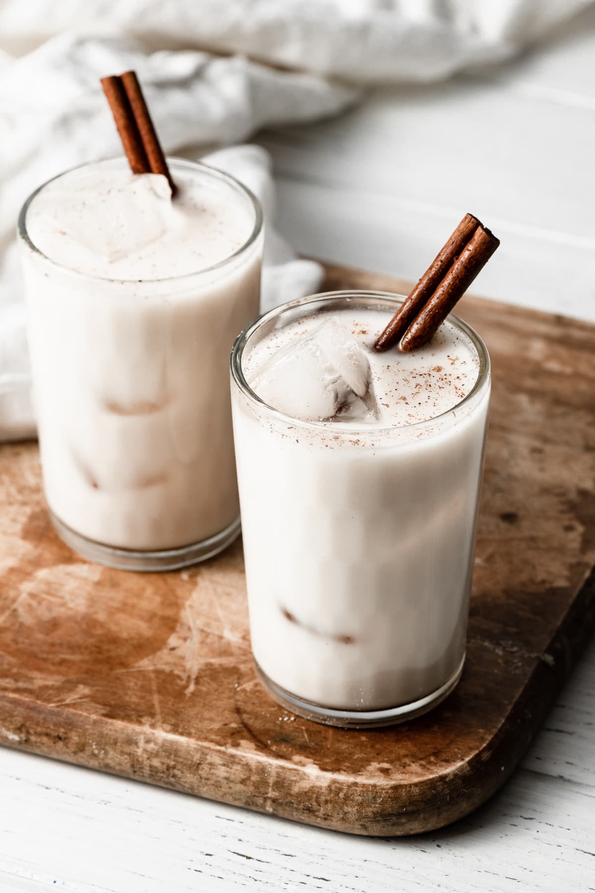 Horchata Horchata in two glass cups with ice cubes and a cinnamon stick. Cups are set over a wooden cutting board.
