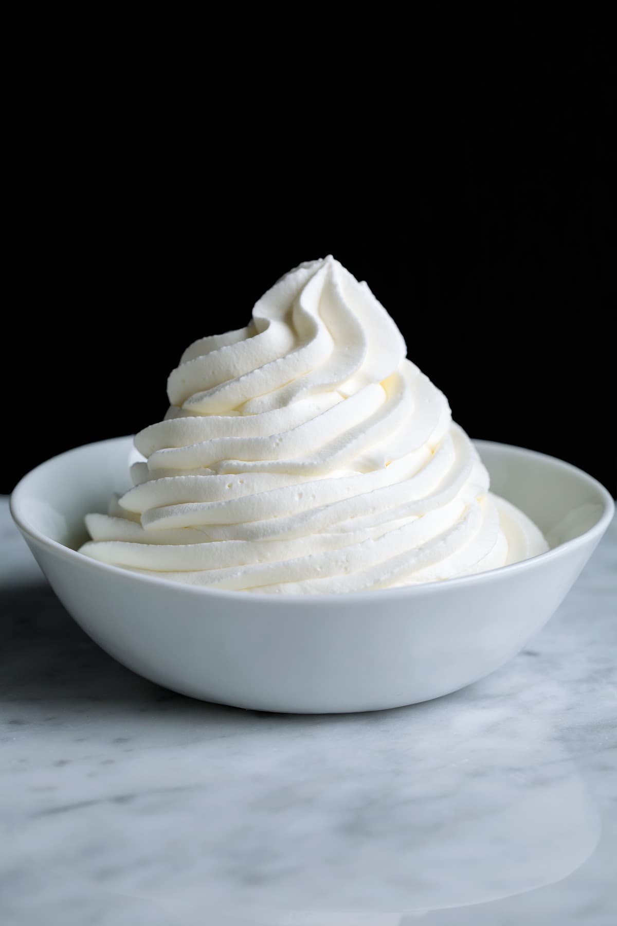 How to Make Whipped Cream Piped mound of whipped cream in a white bowl set over a marble surface with a black background.