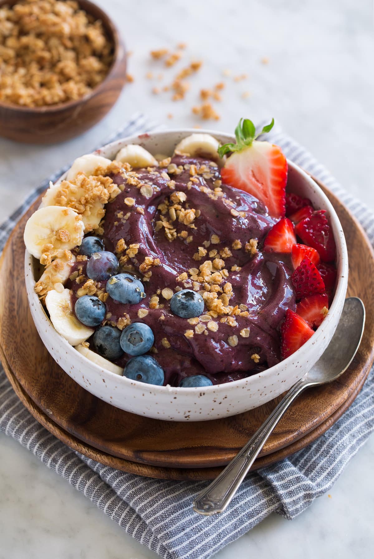 Acai Bowl Thick acai bowl topped with strawberries, blueberries, bananas, granola and honey. Bowl is set over a wooden plate and a grey striped napkin.
