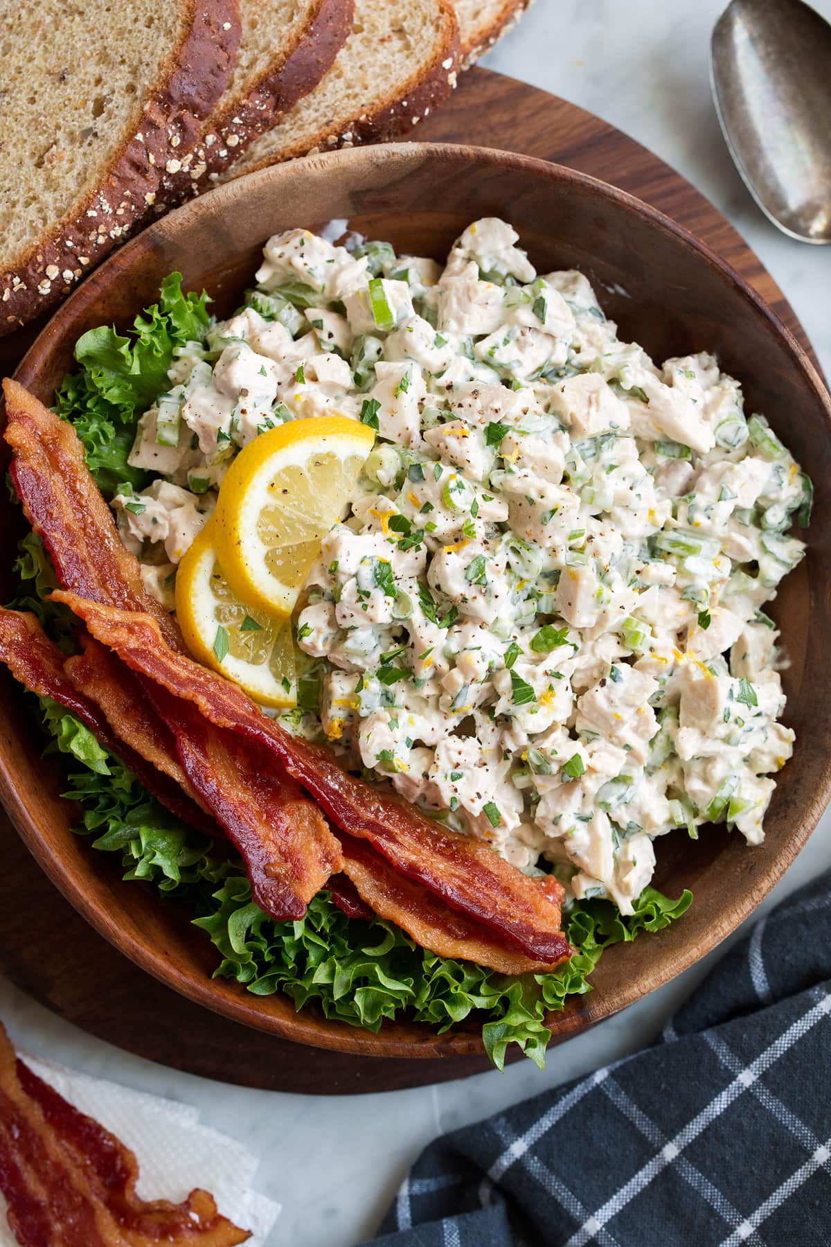 Chicken Salad Chicken salad in a wooden bowl with a side of bacon and lettuce. It is garnished with lemon slices and there are slices of bread to the side of the bowl.