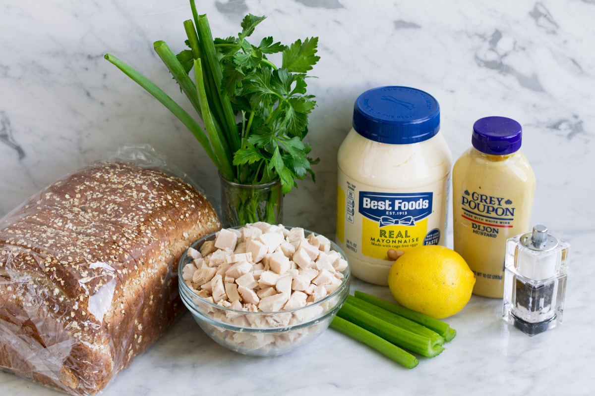 The Best Chicken Salad! Chicken salad ingredients on a marble surface including cooked chicken breast pieces, celery, lemon, mayonnaise, dijon mustard, parsley, green onion, salt and pepper and bread for serving.