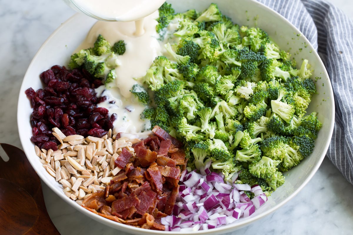Perfect Broccoli Salad! A classic must have recipe! Made with crisp pieces of tiny broccoli florets, salty bacon, sweet dried cranberries, bright red onion, nutty toasted almonds and a creamy rich dressing. #broccoli #salad #summer #sidedish #recipe Pouring dressing over broccoli salad ingredients in a large white bowl.