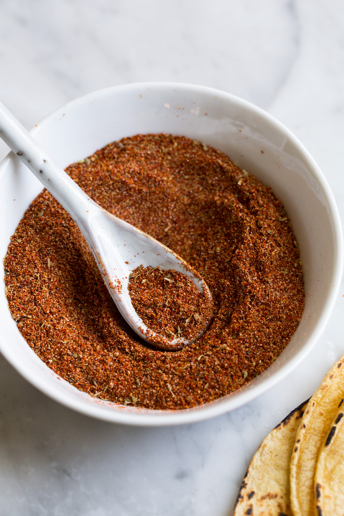 Homemade Taco Seasoning Blended taco seasoning in a small white ceramic bowl with a white ceramic spoon scooping up the seasoning.