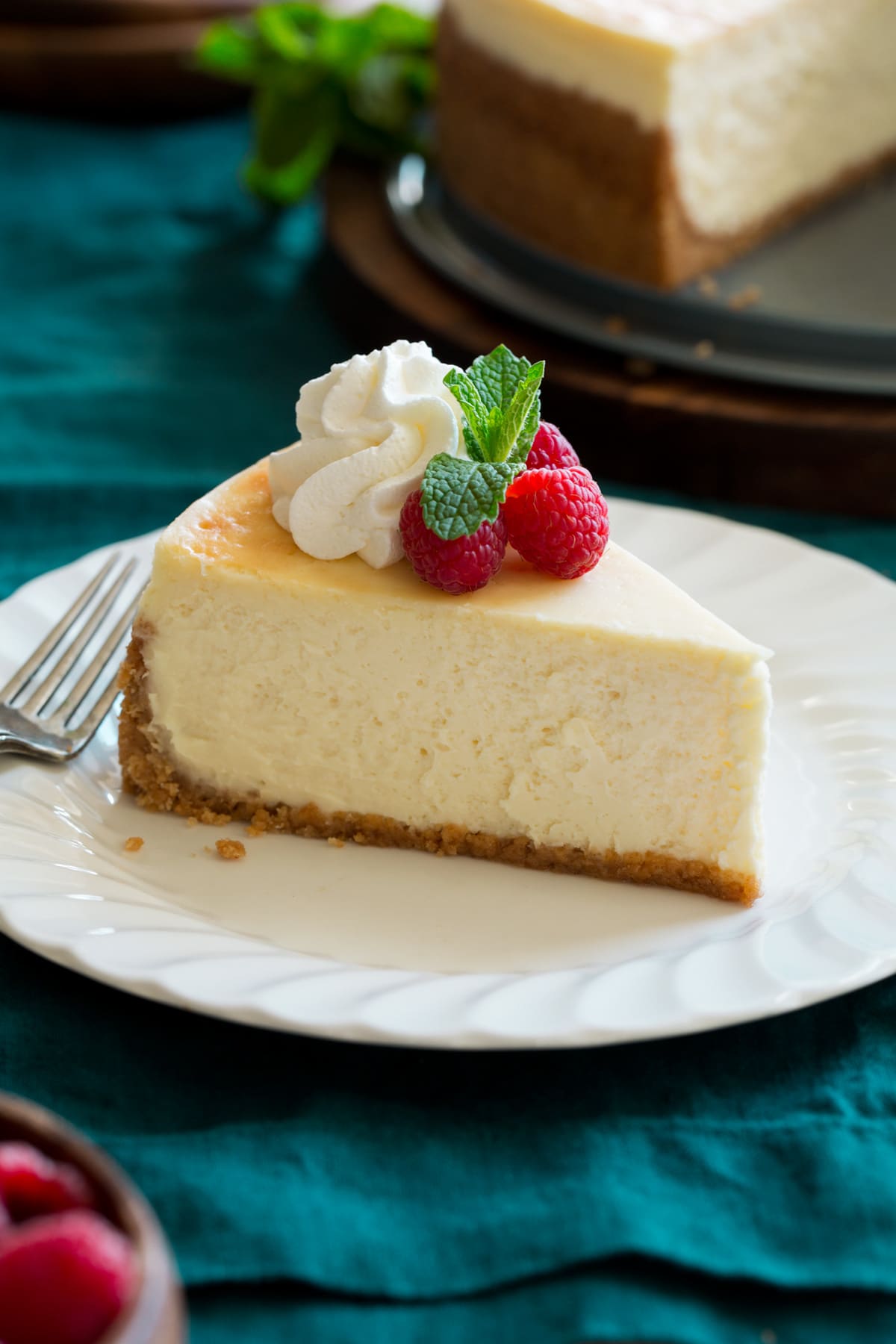 The BEST Cheesecake Recipe! Slice of cheesecake on a white dessert plate sitting over a turquoise cloth. Cheesecake is decorated with whipped cream, raspberries and mint.