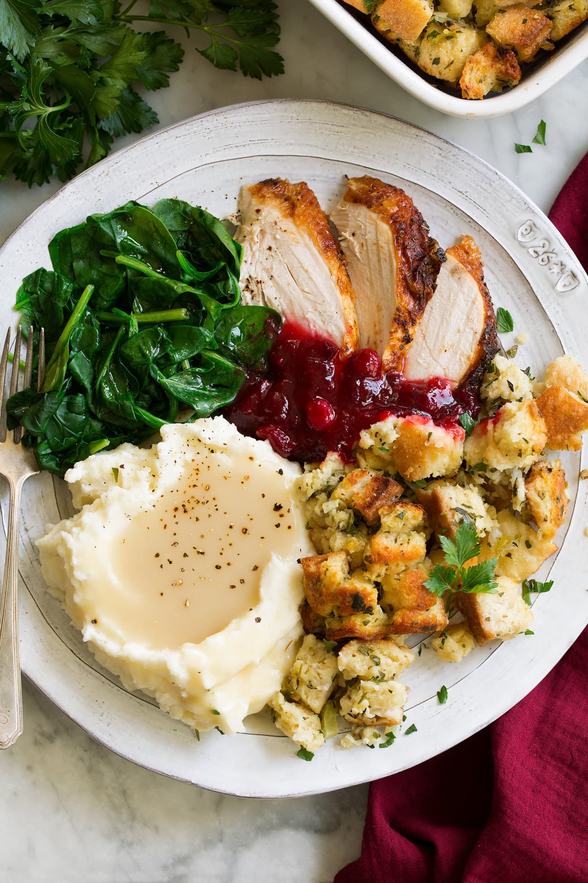 Stuffing Recipe - this is my FAVORITE stuffing! It's perfectly moistened in the center, browned and crisp on the edges and packed with butter and herb flavor. A fall staple! #stuffing #thanksgiving #sidedish #recipe Stuffing on a large dinner plate with a side of mashed potatoes and gravy, turkey and steamed spinach.