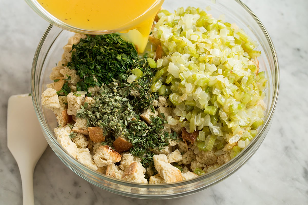 Stuffing Recipe - this is my FAVORITE stuffing! It's perfectly moistened in the center, browned and crisp on the edges and packed with butter and herb flavor. A fall staple! #stuffing #thanksgiving #sidedish #recipe Showing how to make stuffing. Pouring broth mixture over dried bread cubes, celery mixture and herbs in a glass mixing bowl.
