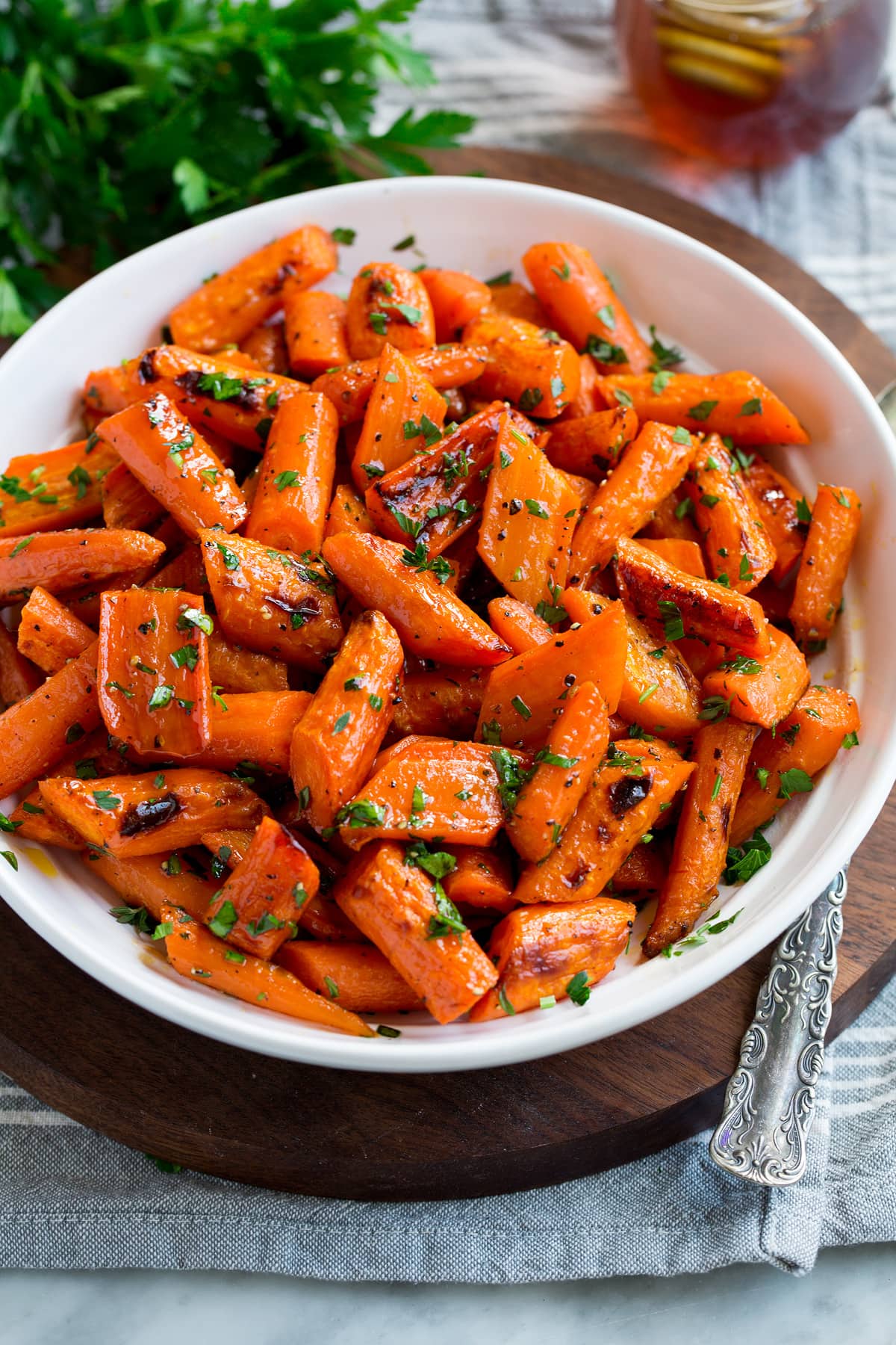 Roasted Carrots Photo: Roasted carrots in a white serving bowl. Carrots are garnished with parsley with a bunch in the background. Bowl is resting on a wooden platter and a grey cloth.