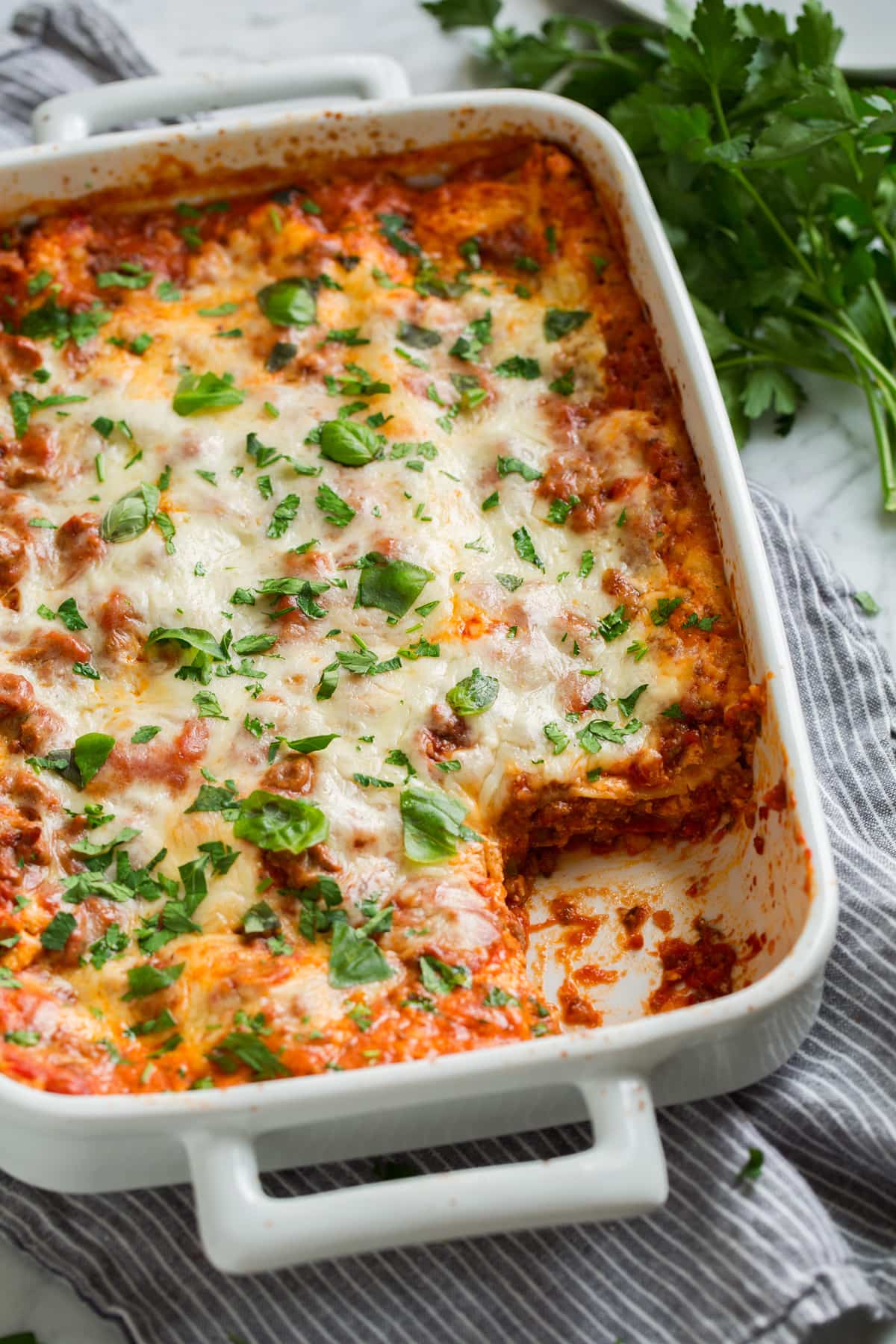 Easy Lasagna Recipe Finished lasagna in a white casserole dish with one slice scooped out. Dish shown tilted at an angle with fresh parsley in the background.
