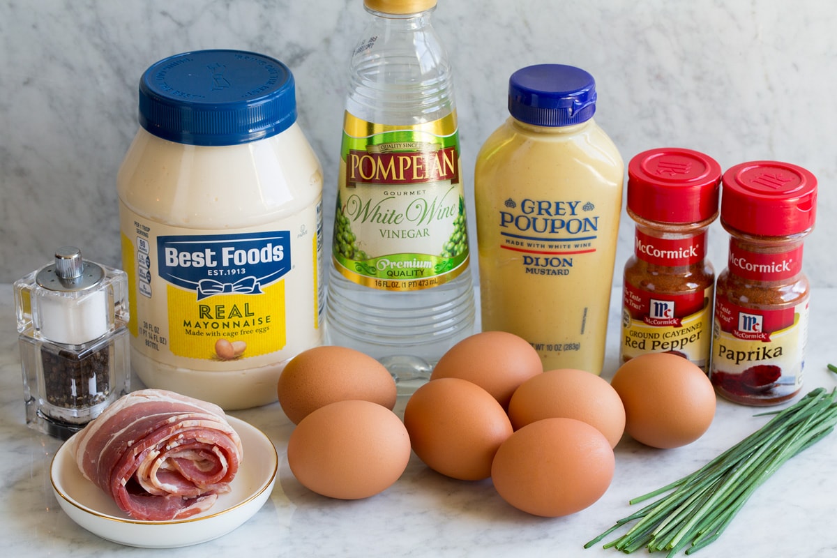 Best Deviled Eggs Image showing ingredients that go into deviled eggs including mayonnaise, dijon mustard, white wine vinegar, eggs, chives, cayenne pepper, paprika, chives and bacon.