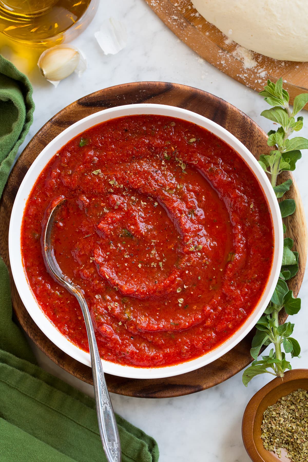 Homemade Pizza Sauce Overhead image of pizza sauce in a white bowl with a spoon in the sauce. Sauce is resting on a wooden plate over a marble surface.