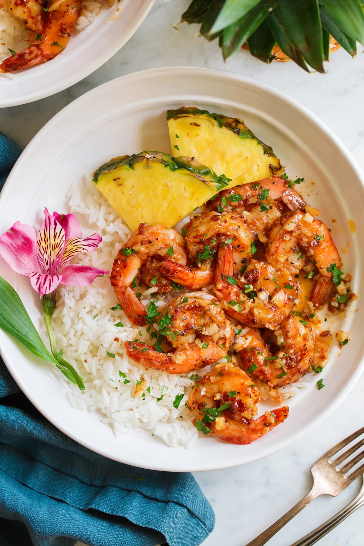 Hawaiian Garlic Shrimp Garlic shrimp in a shallow white bowl with rice, pineapple and a fresh tropical flower for decoration.