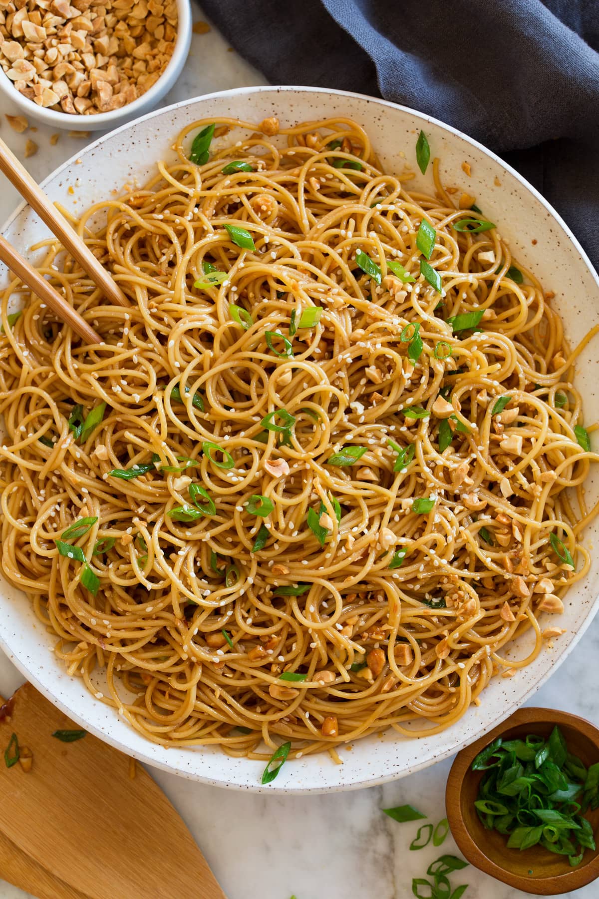 Sesame Noodles Photo: Bowl of sesame noodles topped with peanuts, sliced green onions and sesame seeds. Shown overhead in a white bowl with a dark napkin underneath and chopsticks and serving spoons to the side.
