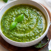Bowl full of fresh homemade pesto. Bowl is resting on a wooden platter. Parmesan, fresh basil and pine nuts are shown in the background.