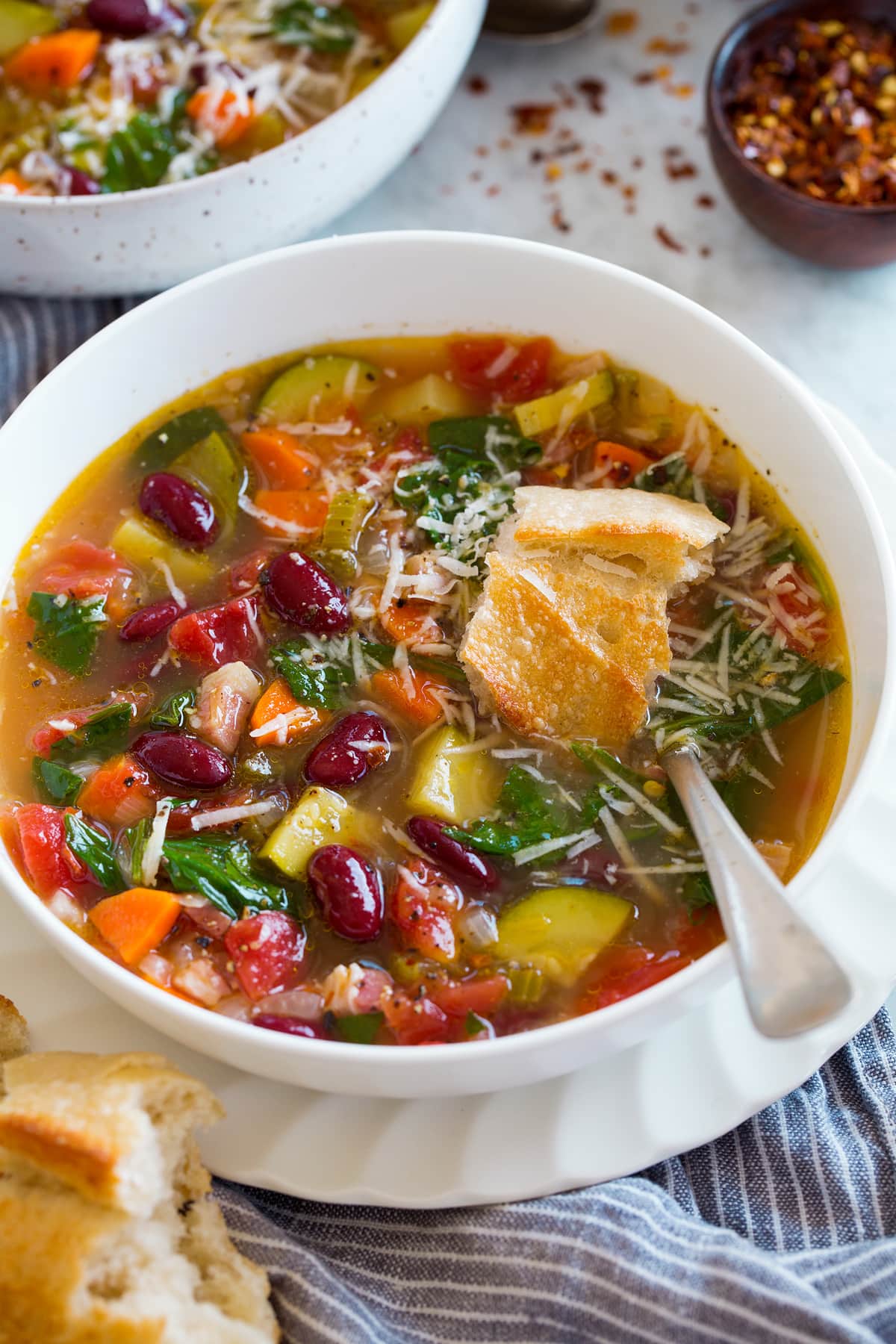 Minestrone Soup Image of serving of minestrone soup in a white bowl set over white plate. Soup is garnished with parmesan and topped with bread.