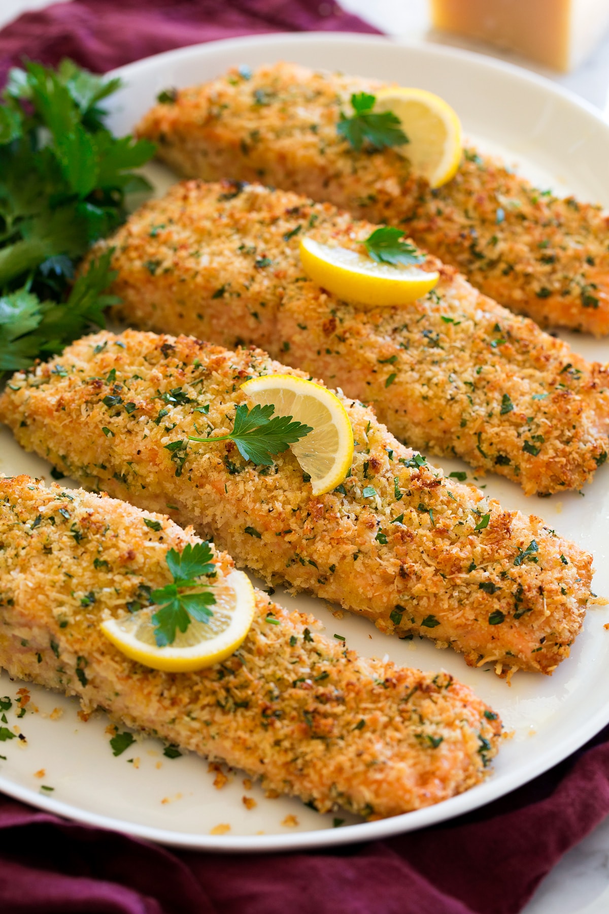 Baked Parmesan Crusted Salmon Four fillets of baked Parmesan Crusted Salmon shown on a white oval serving platter garnished with lemon slices and parsley.