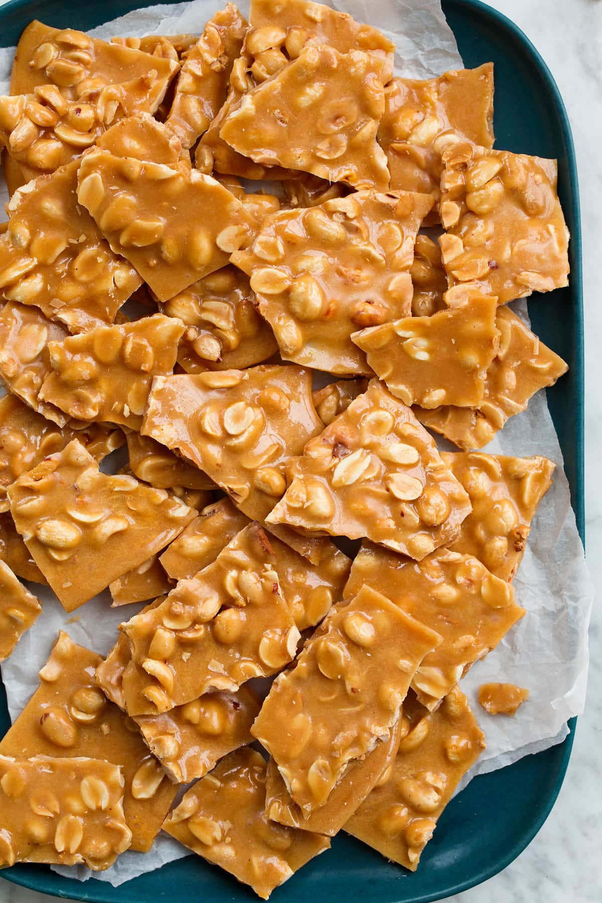 Peanut Brittle Close up overhead image of peanut brittle stacked on a blue platter with parchment paper.