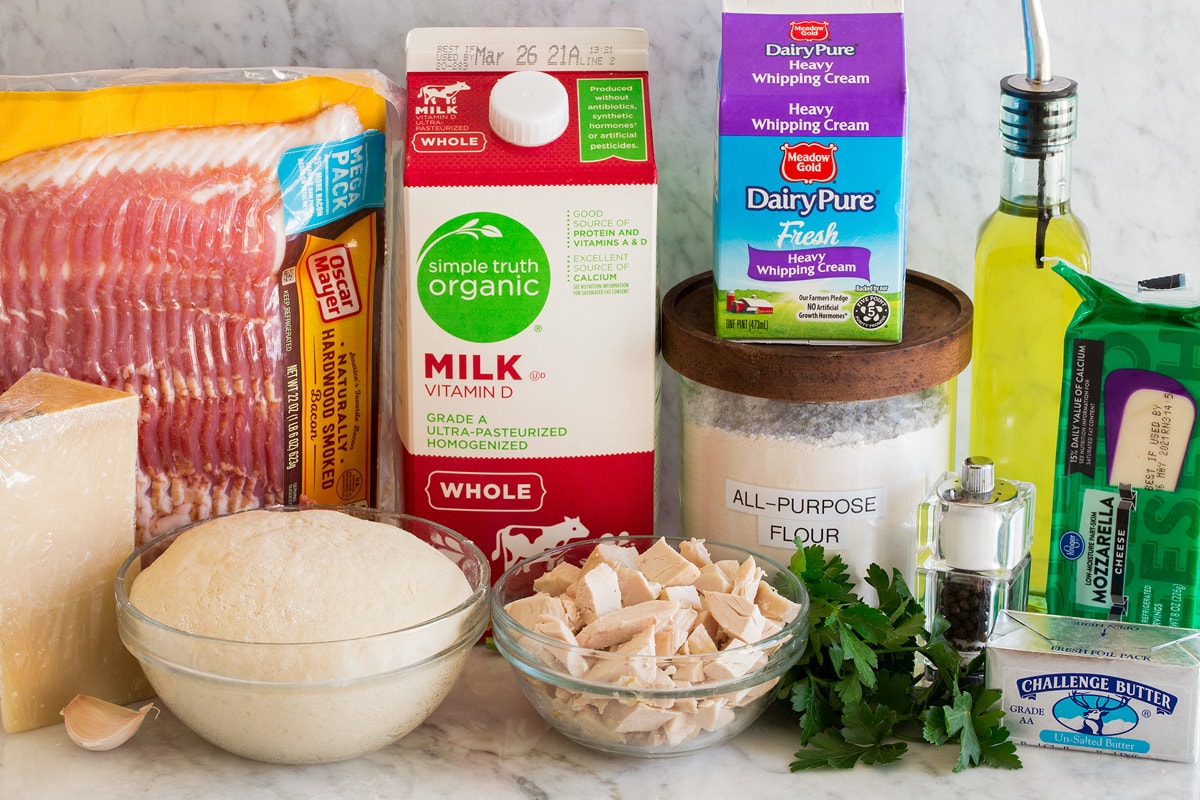 Chicken Alfredo Pizza Photo of ingredients for making chicken alfredo pizza. Includes bacon, parmesan, pizza dough, garlic, chicken, parsley, flour, heavy cream, milk, olive oil, mozzarella cheese and butter.