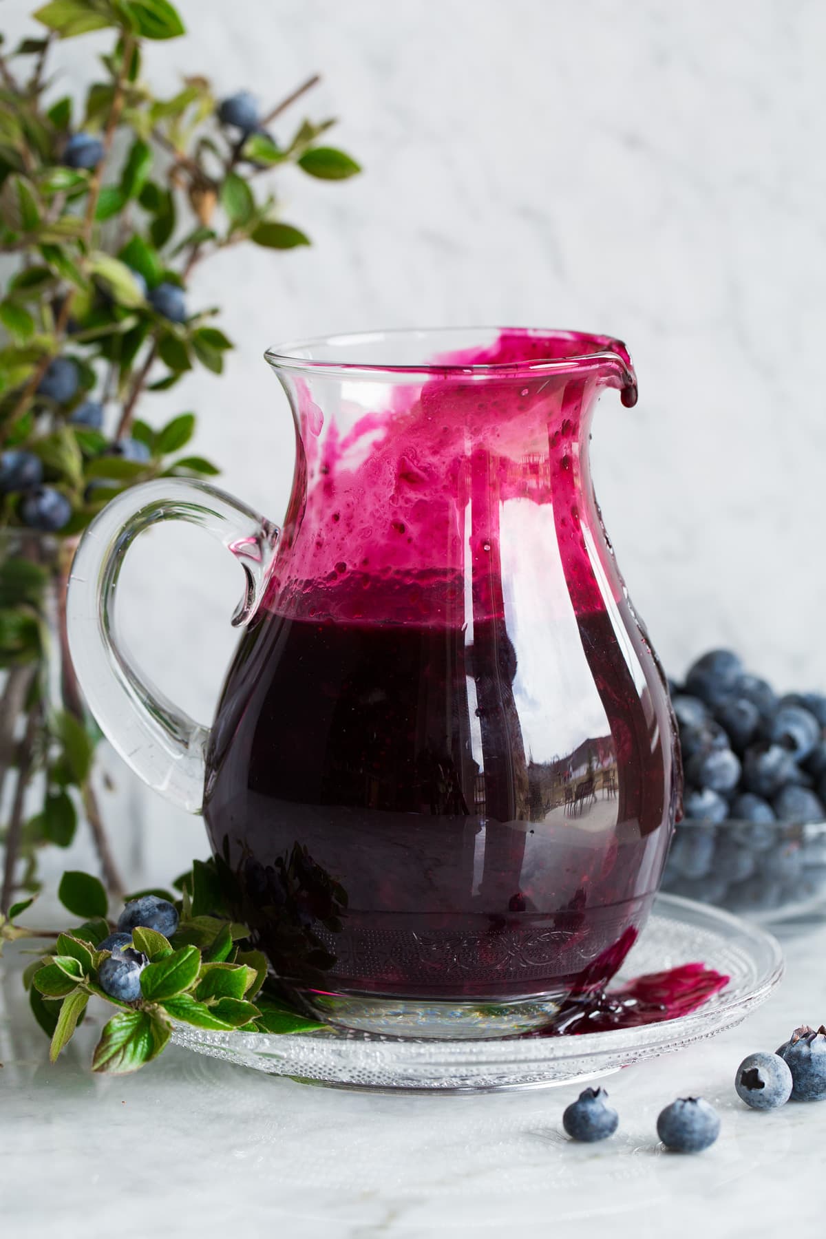 Blueberry Syrup Blueberry syrup in a glass pitcher set over a glass plate.