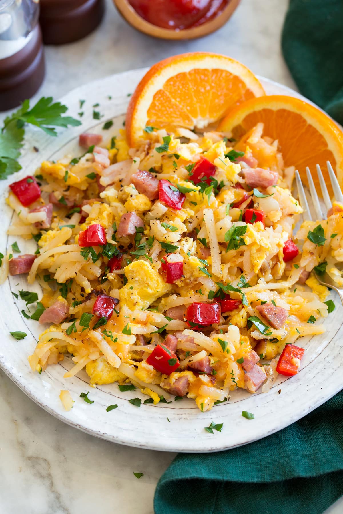 Breakfast Hash Photo: Serving of breakfast hash on a white ceramic plate with oranges to the side. Hash is made with shredded hash browns, ham, eggs, bell pepper, parsley and onion.