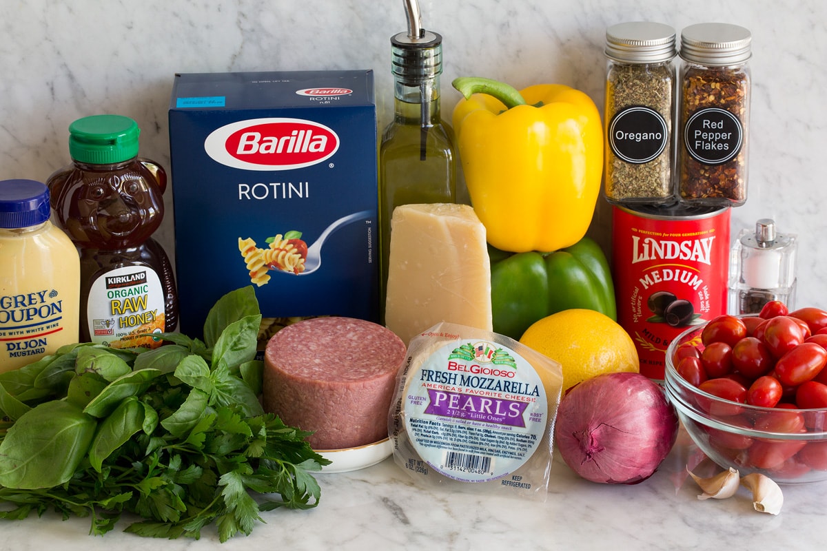 Italian Pasta Salad Photo: Ingredients displayed that are used to make Italian pasta salad. Includes rotini pasta, bell peppers, onion, lemon, parmesan, mozzarella, tomatoes, olives, herbs, garlic, salami, honey and dijon.