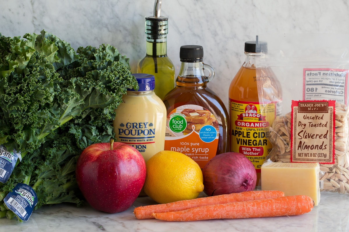 Kale Salad Photo showing ingredients used to make kale salad. Includes kale, apples, lemon, red onion, carrots, white cheddar, maple syrup, apple cider vinegar, dijon mustard, olive oil and slivered almonds.