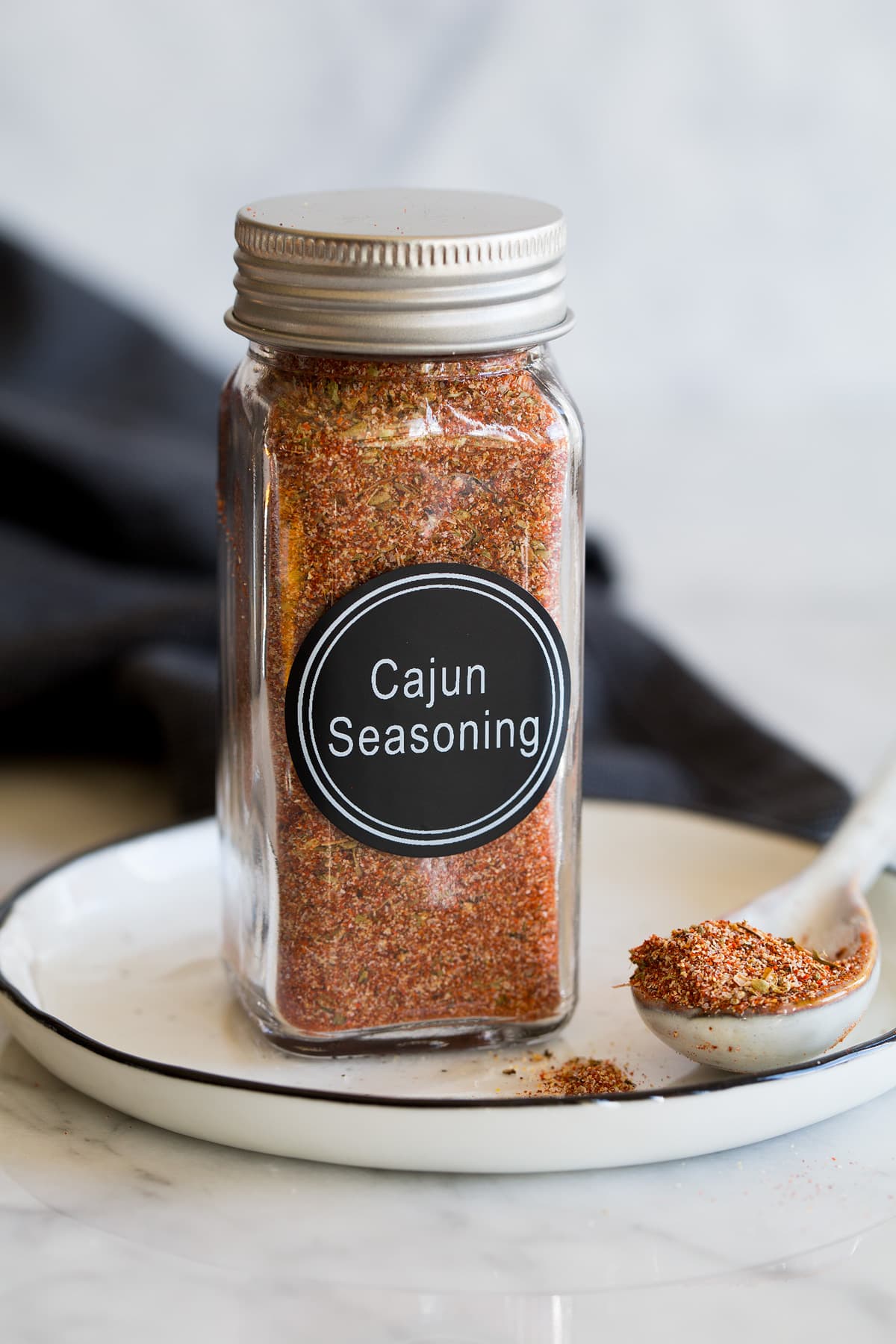Homemade Cajun Seasoning Photo: Homemade cajun seasoning blend in a glass spice jar on a small plate with a spoon to the side and a dark napkin in the background.
