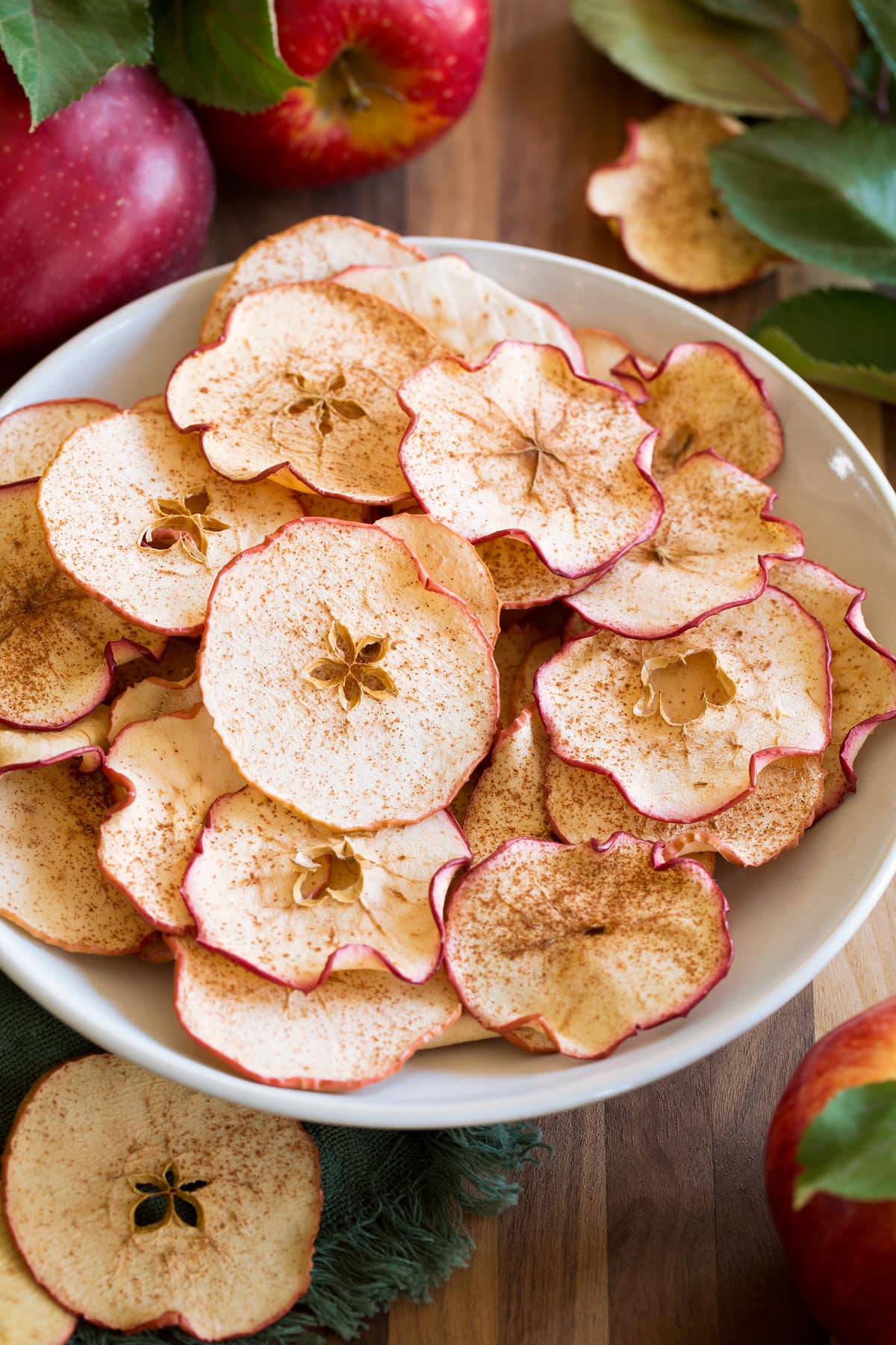 Baked Apple Chips Apple chips shown in a white bowl on a wooden tabletop with fresh apples and leaves surrounding them.