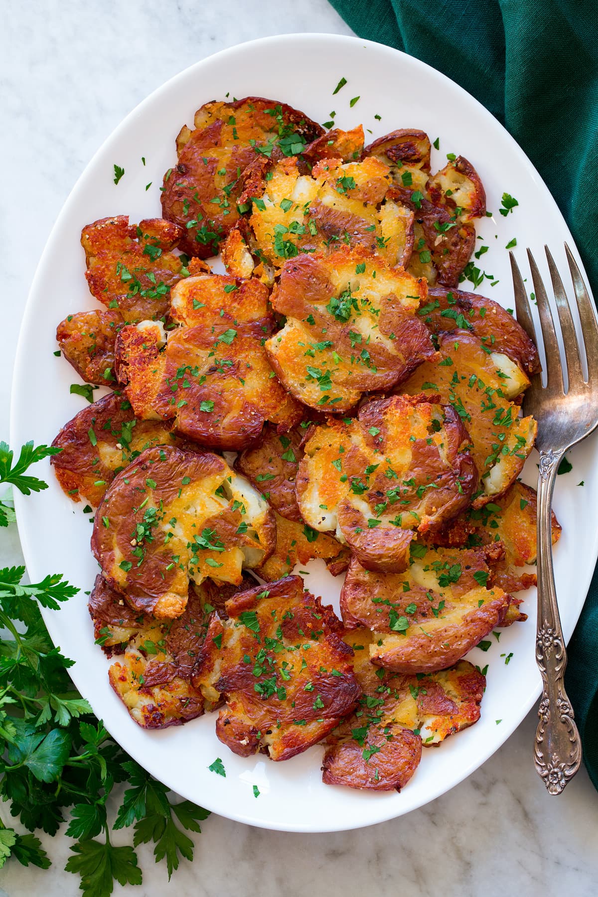 Smashed Red Potatoes Smashed red potatoes shown piled on a white oval platter. They are shown from above on a white marble surface with a green cloth and parsley to the sides.