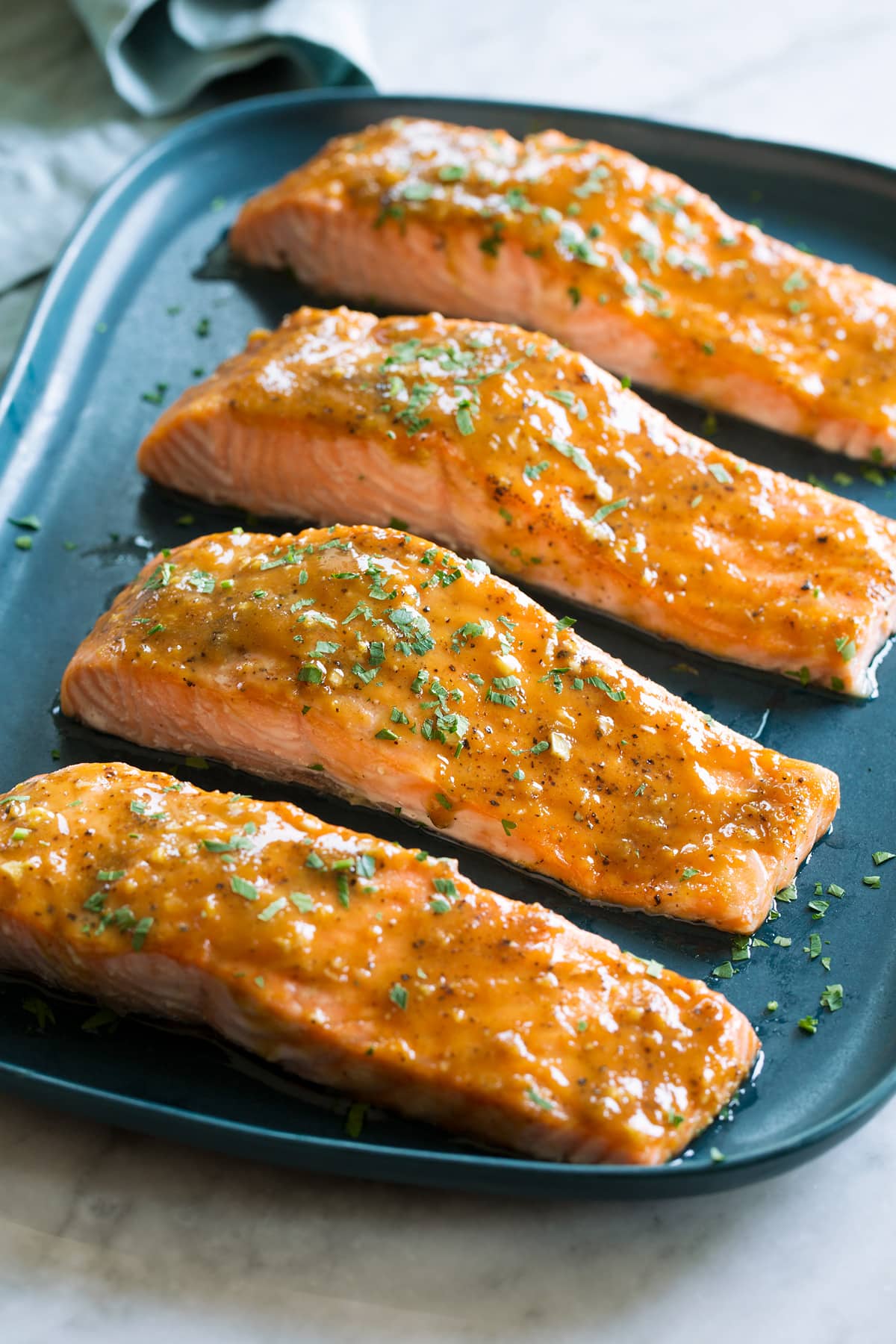 Brown Sugar Glazed Salmon Photo of four salmon fillets coated with a brown sugar glaze resting on a blue rectangular platter on a white marble surface.
