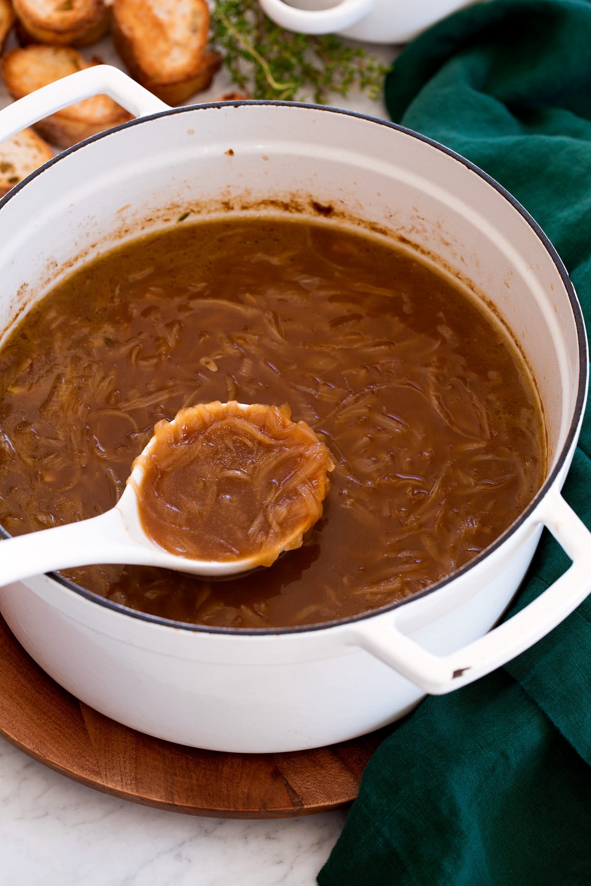 French Onion Soup Large white pot filled with french onion soup. A ladle is scooping soup and pot is resting on a wooden platter with a green cloth.
