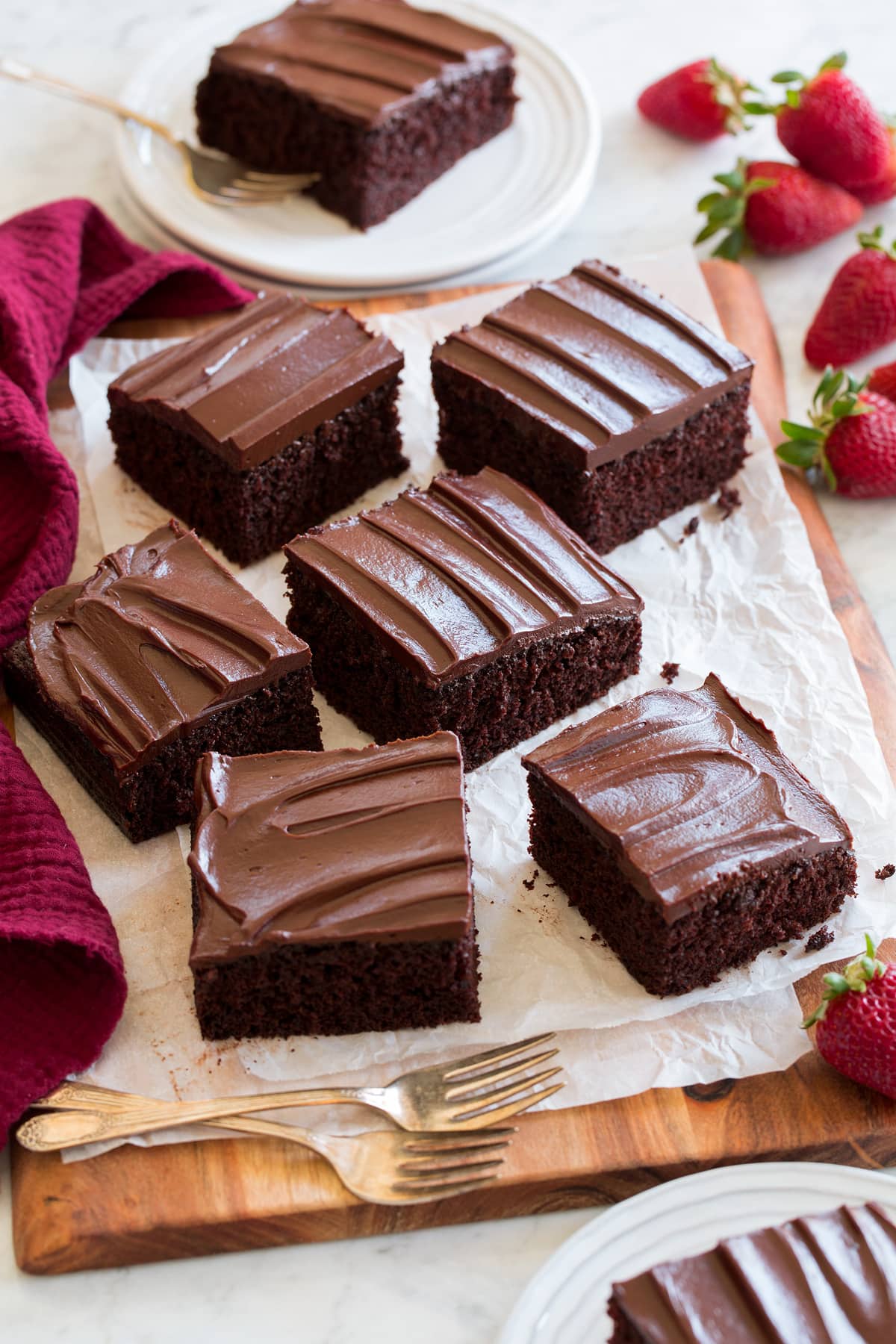 Easy Chocolate Cake Square slices of homemade chocolate cake shown on a wooden serving board with parchment paper. Strawberries are shown as a serving suggestion to the side.
