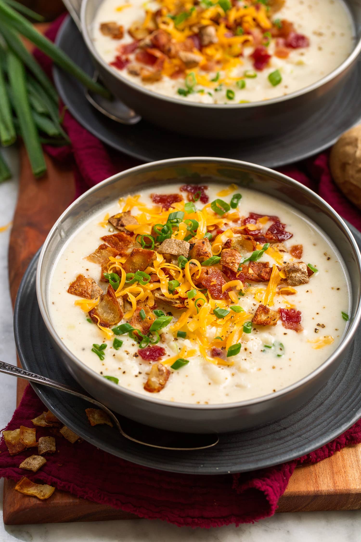 Baked Potato Soup Serving of baked potato soup in a gray bowl over a gray plate. It is topped with cheddar cheese, fried potato skins, bacon and green onions.
