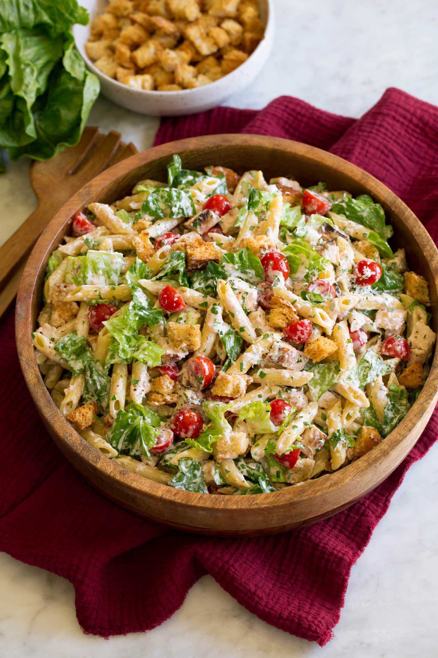 Chicken Caesar Pasta Salad Chicken caesar pasta salad shown in a brown wooden serving bowl over a red cloth on a white marble surface.