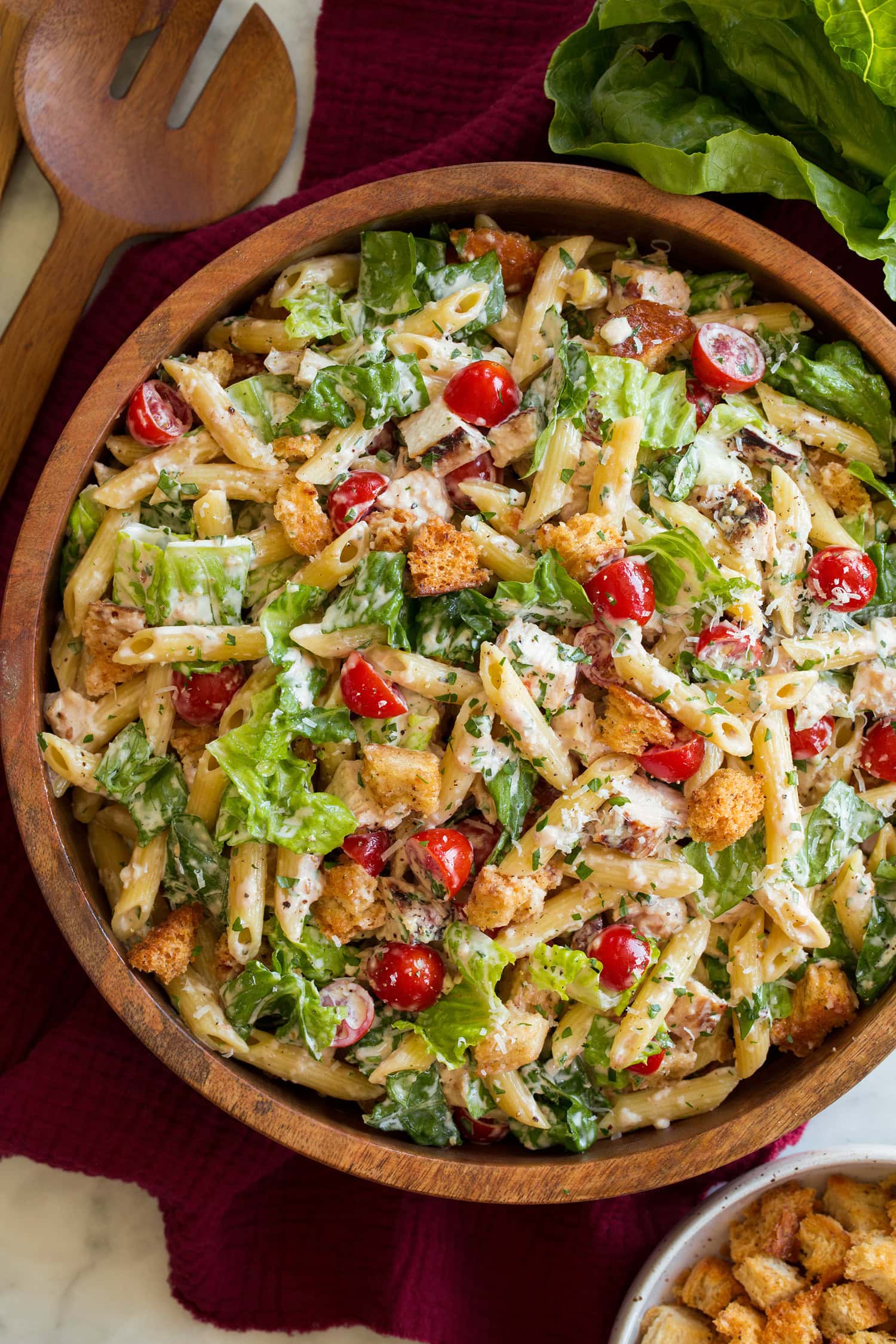 Chicken Caesar Pasta Salad Grilled chicken caesar salad shown from overhead in a wooden bowl with romaine lettuce and wooden serving spoons to the side.