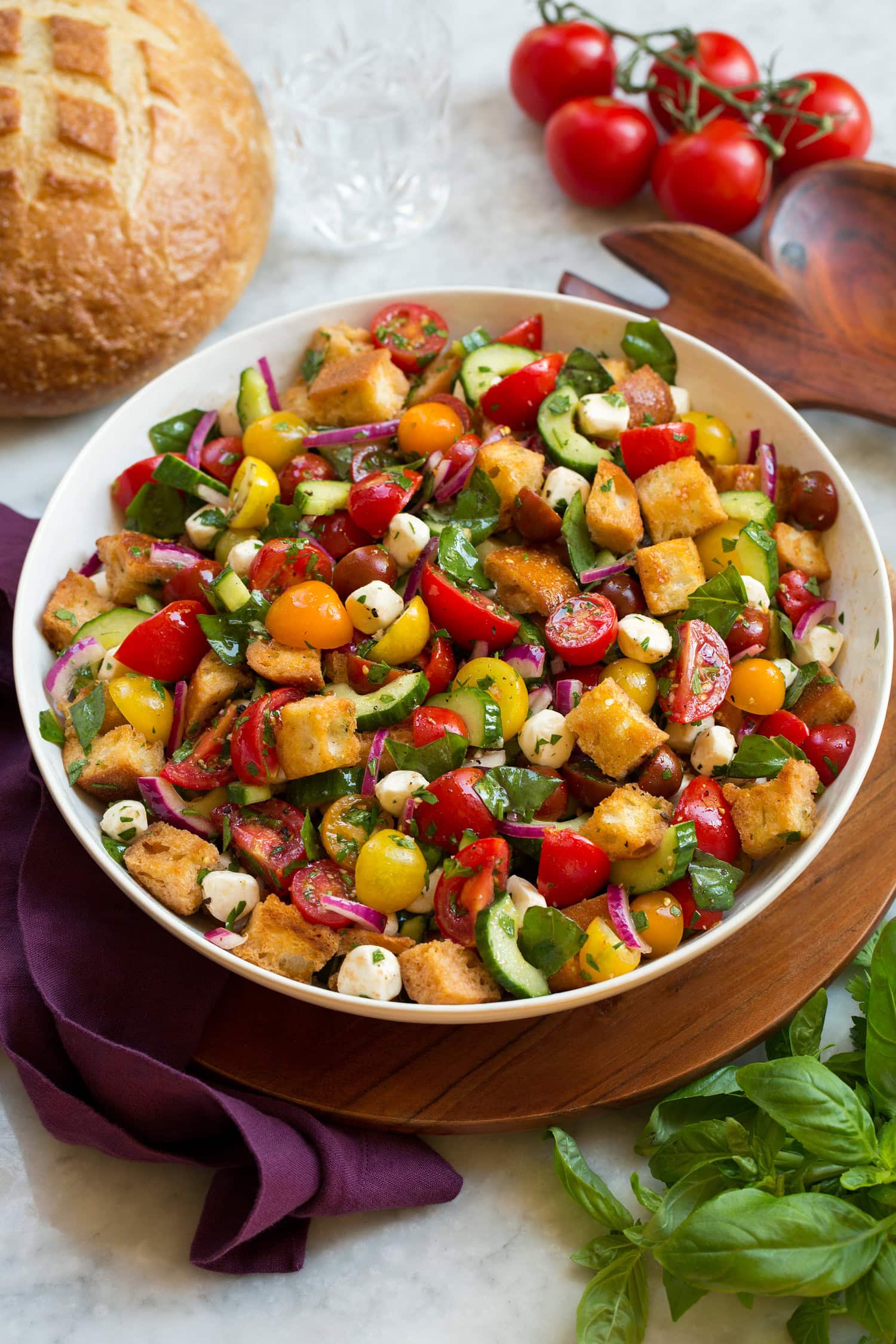 Panzanella Salad Panzanella salad in a white bowl over a wooden tray with a purple cloth and fresh basil and tomatoes shown to the sides.