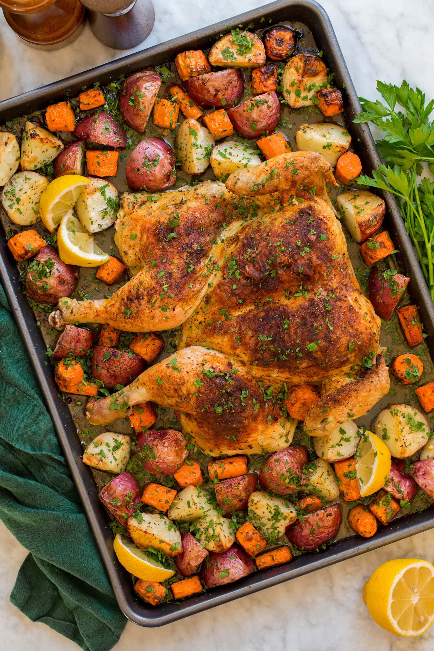 Spatchcock Chicken Spatchcock chicken with roasted vegetables on a dark rimmed baking sheet with a green cloth to the side.