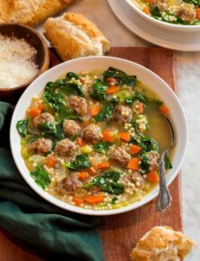Italian wedding soup in a serving bowl with baguette bread and parmesan shown for serving.