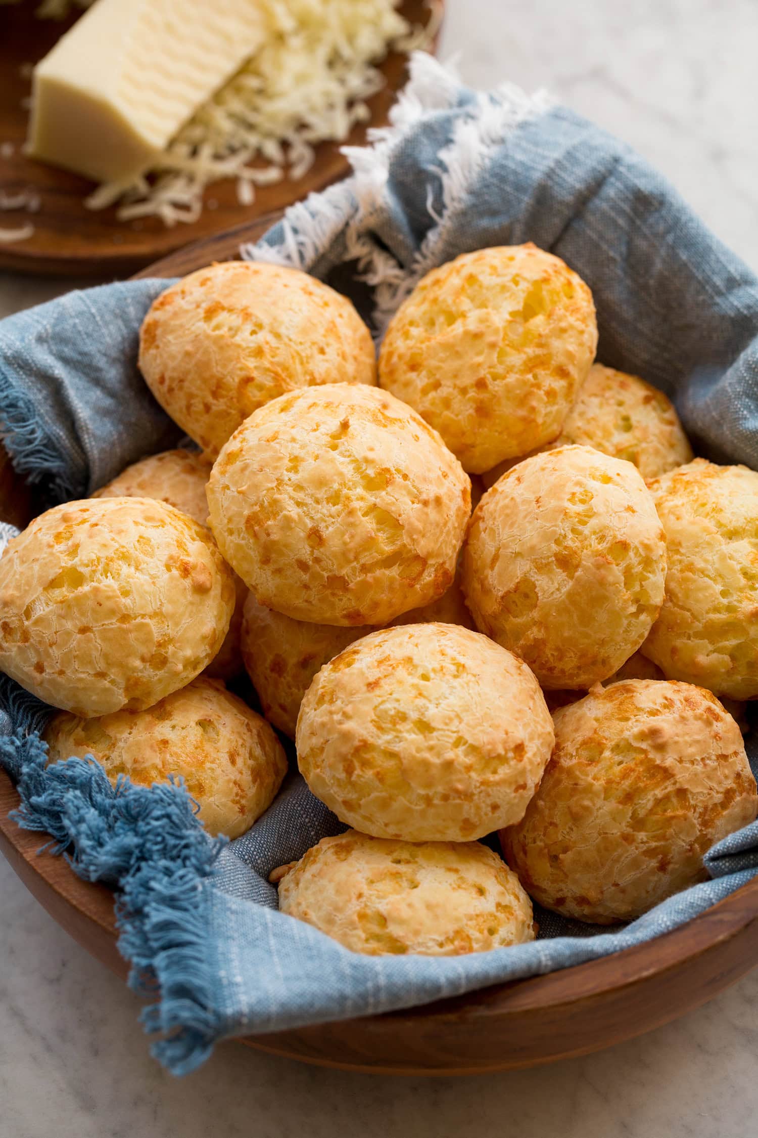Brazilian Cheese Bread (Pão de Queijo) Homemade Brazilian cheese bread in a wooden bowl with a blue cloth underneath the bread. Grated cheese is in the background.